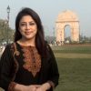 Smiling woman in a black embroidered kurta posing outdoors with India Gate in the background.