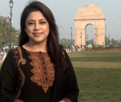 Smiling woman in a black embroidered kurta posing outdoors with India Gate in the background.