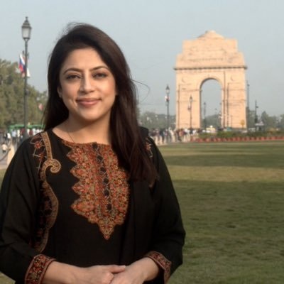 Smiling woman in a black embroidered kurta posing outdoors with India Gate in the background.