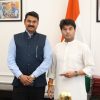 Two men in formal attire pose in an official room, with an Indian flag in the background.