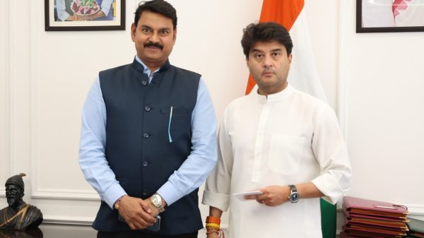 Two men in formal attire pose in an official room, with an Indian flag in the background.
