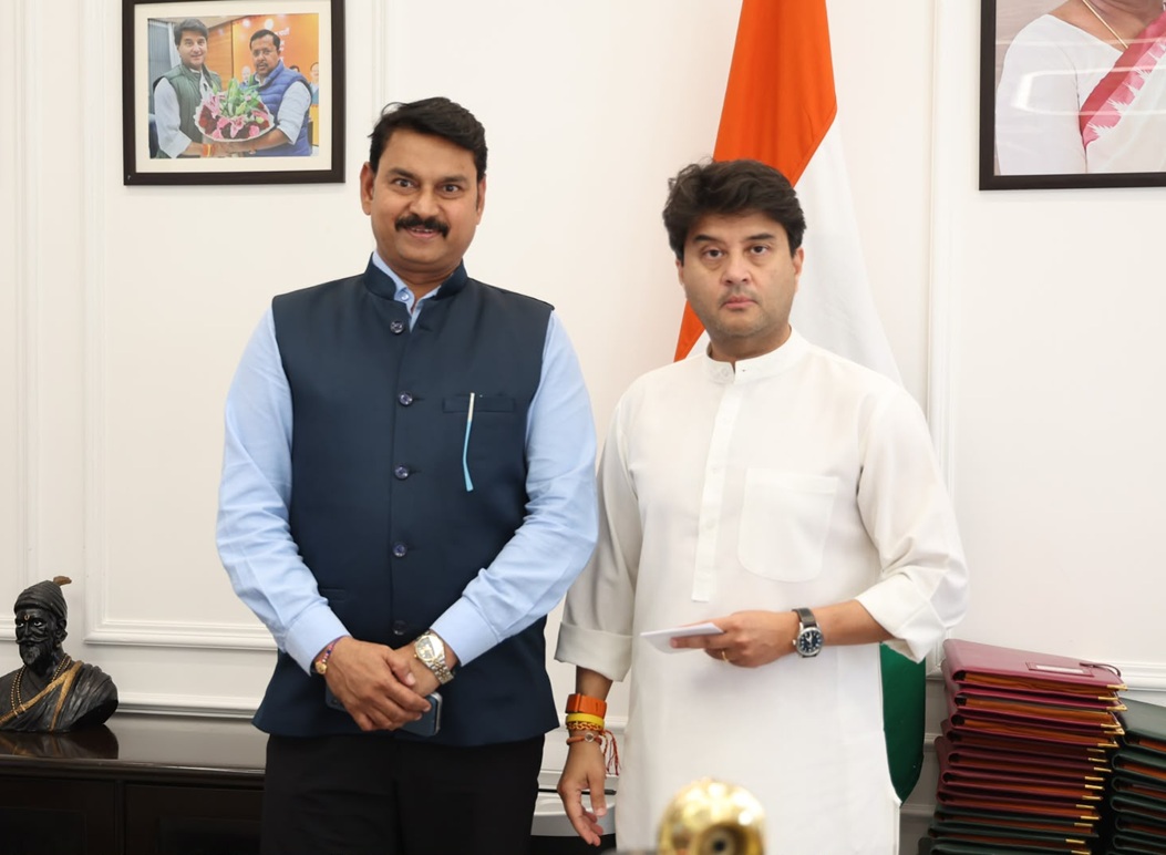 Two men in formal attire pose in an official room, with an Indian flag in the background.