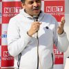 Man speaking into a handheld microphone at a public event, wearing a light gray zip hoodie with a backdrop of red-and-white NBT banners.