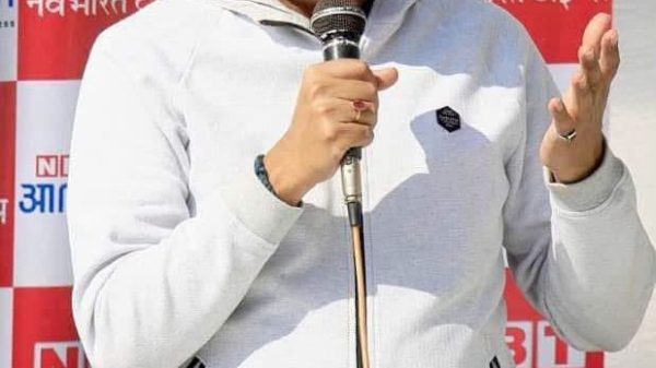 Man speaking into a handheld microphone at a public event, wearing a light gray zip hoodie with a backdrop of red-and-white NBT banners.