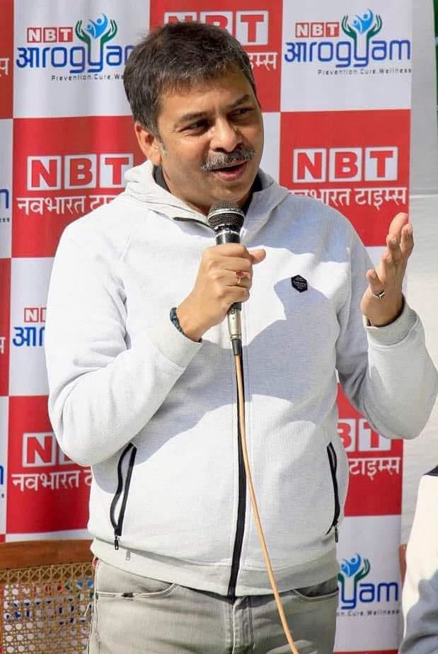 Man speaking into a handheld microphone at a public event, wearing a light gray zip hoodie with a backdrop of red-and-white NBT banners.