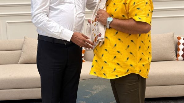 Two men indoors; one in a white dress shirt hands a patterned scarf to the other in a bright yellow shirt, both smiling.