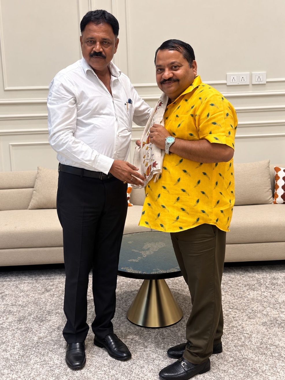 Two men indoors; one in a white dress shirt hands a patterned scarf to the other in a bright yellow shirt, both smiling.