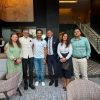 Six people stand together in a modern hotel lobby posing for a group photo in smart casual and business attire.