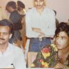 Two men sit at a social gathering; one in a light shirt with a mustache, the other in a bright floral shirt, with guests in the background in a party setting.