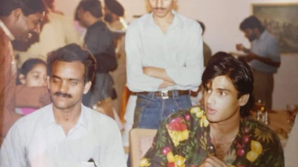 Two men sit at a social gathering; one in a light shirt with a mustache, the other in a bright floral shirt, with guests in the background in a party setting.