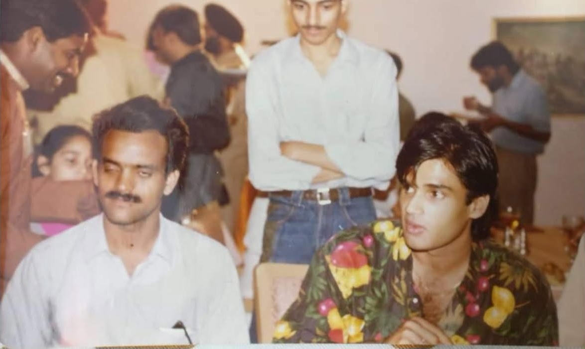 Two men sit at a social gathering; one in a light shirt with a mustache, the other in a bright floral shirt, with guests in the background in a party setting.