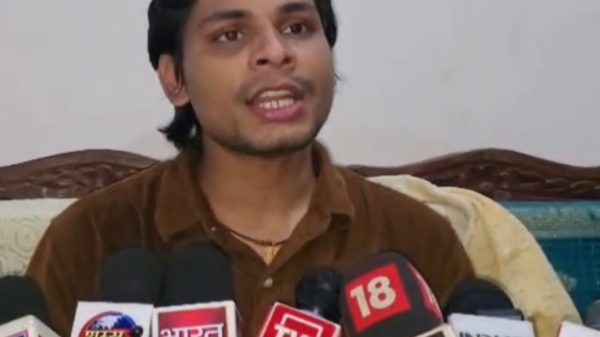 Man in a brown shirt speaks at a press conference, microphones from multiple news outlets in front of him.