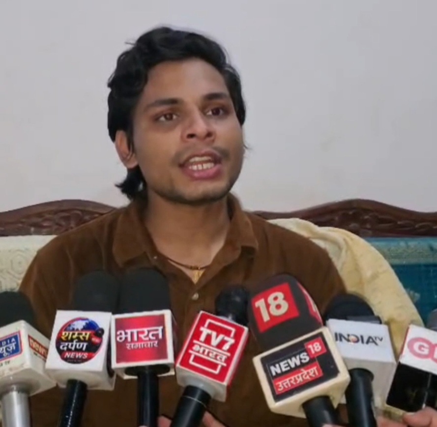 Man in a brown shirt speaks at a press conference, microphones from multiple news outlets in front of him.