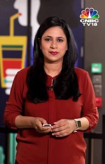 Female news anchor in a red blouse stands in a studio, holding a small device, CNBC TV18 logo in the top-right corner.