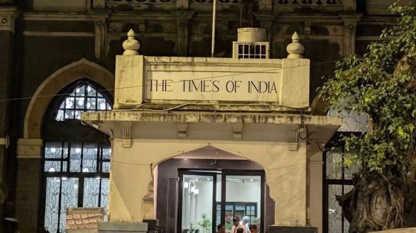 Night view of The Times of India building entrance with its sign, illuminated doorway, and people inside; a yellow taxi and a red car are in the street in the foreground.