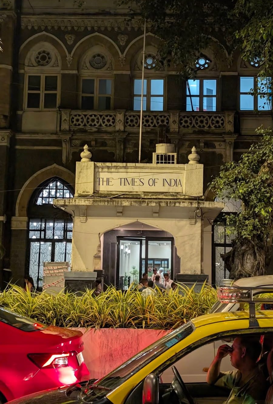 Night view of The Times of India building entrance with its sign, illuminated doorway, and people inside; a yellow taxi and a red car are in the street in the foreground.