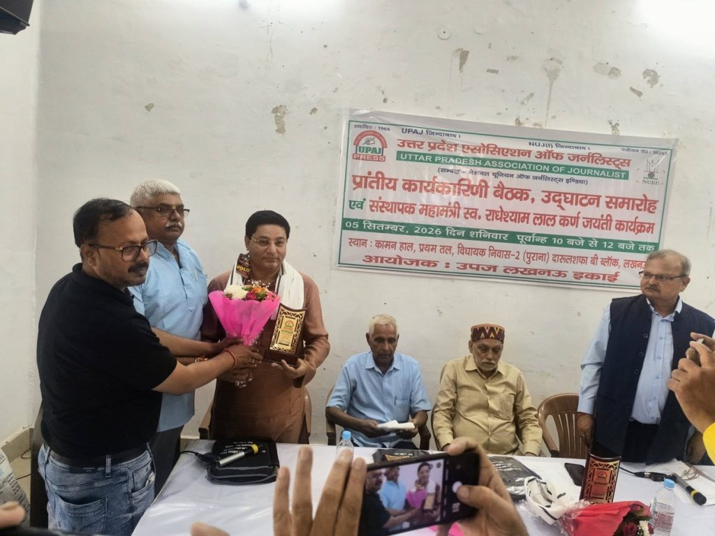 Man presents a bouquet and a plaque to a woman at a ceremony, with three men seated at a table and a Hindi banner on the wall behind them.