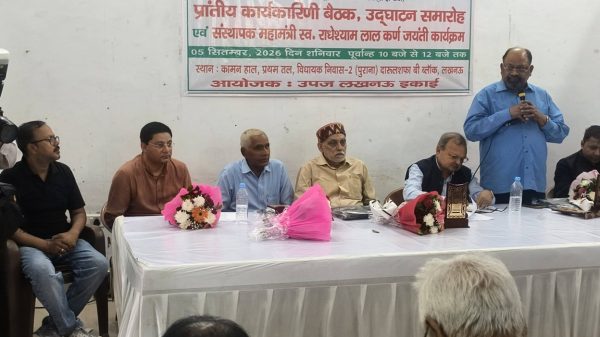 Panel discussion at a press conference with six men seated behind a table, one man standing and speaking into a microphone as flowers sit on the table. A banner in a foreign script hangs on the wall behind them.