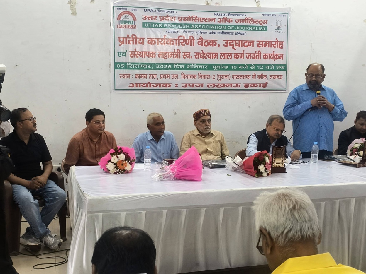Panel discussion at a press conference with six men seated behind a table, one man standing and speaking into a microphone as flowers sit on the table. A banner in a foreign script hangs on the wall behind them.