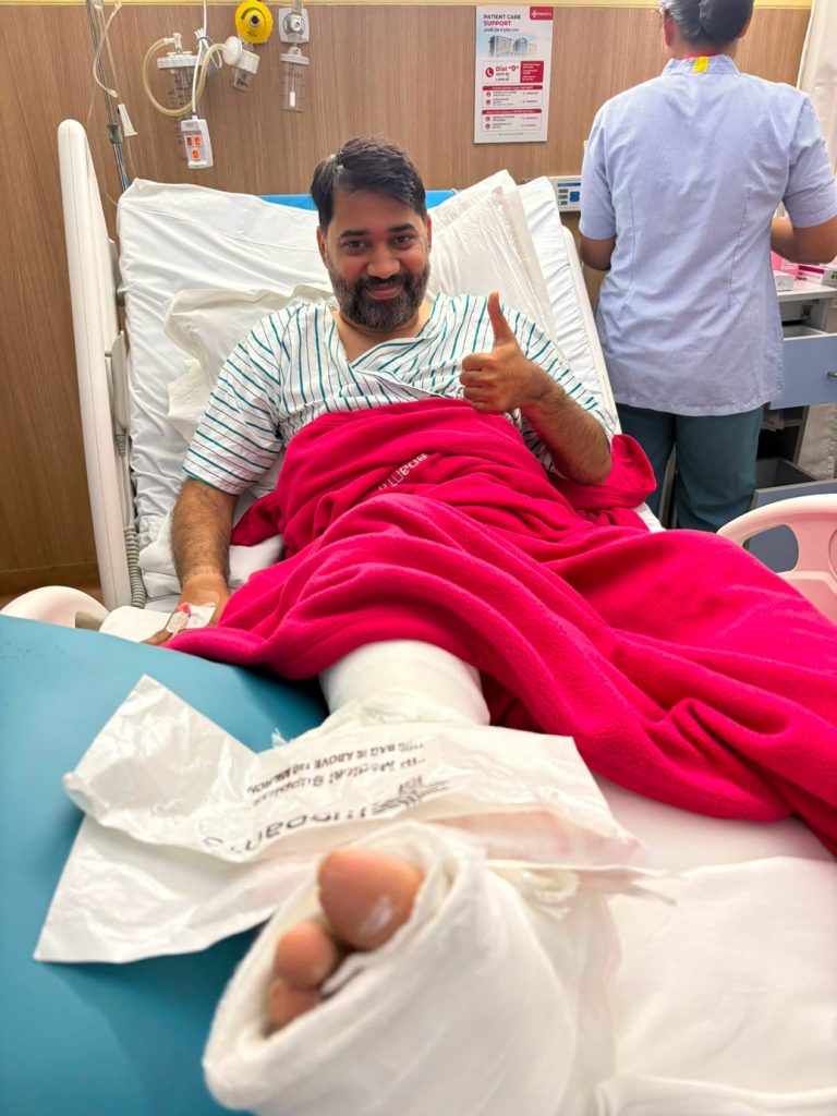 Smiling man in a hospital bed giving a thumbs up, with a pink blanket over his leg in a cast; nurse stands nearby in a blue uniform.