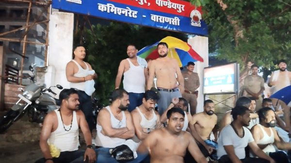 Group of shirtless men sitting on a street at night, with a police officer and a red-blue signboard overhead in Hindi