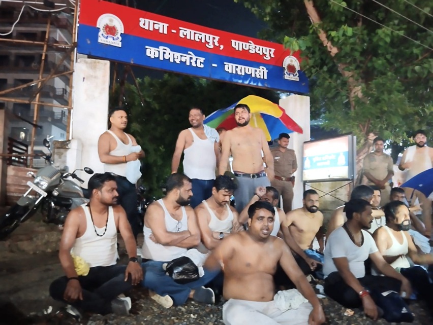 Group of shirtless men sitting on a street at night, with a police officer and a red-blue signboard overhead in Hindi