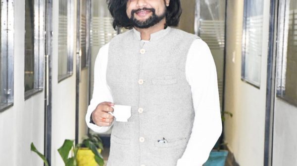 Smiling man with long dark hair and a beard, wearing a gray vest over a white shirt, holding a white cup in a plant-lined hallway.