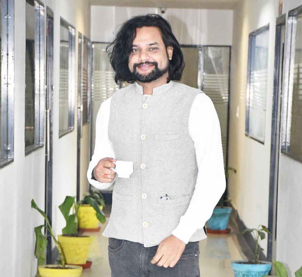 Smiling man with long dark hair and a beard, wearing a gray vest over a white shirt, holding a white cup in a plant-lined hallway.