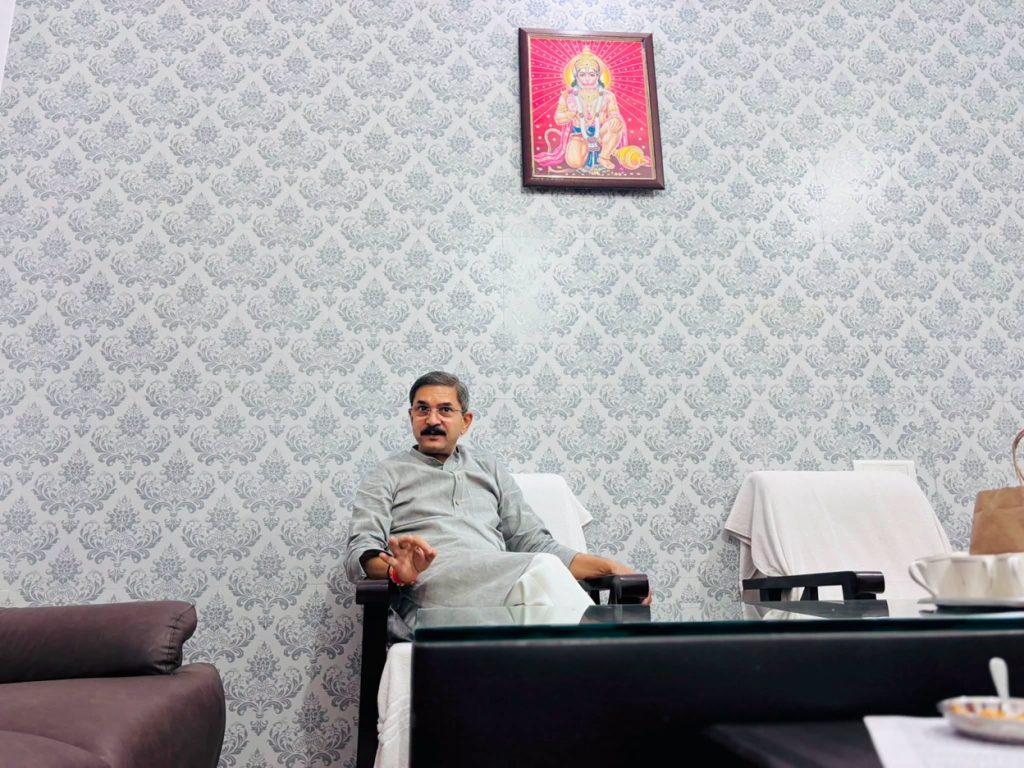 Man sitting in a chair in a room with gray damask wallpaper; a framed pink deity image hangs on the wall above him.