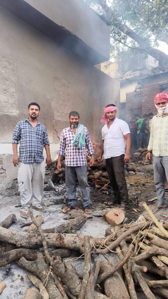 Four men stand among charred logs and rubble outside a smoke-filled, damaged building.