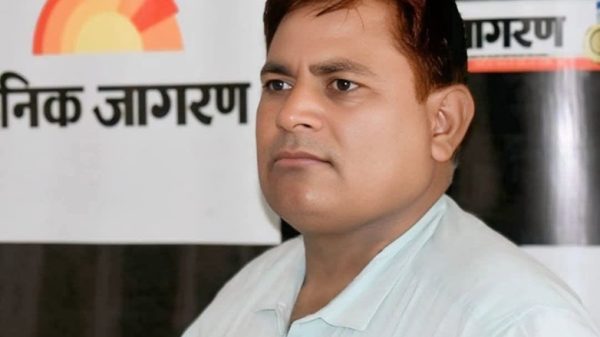 Portrait of a man in a light blue shirt standing in front of a Dainik Jagran banner with a sun logo in the background.