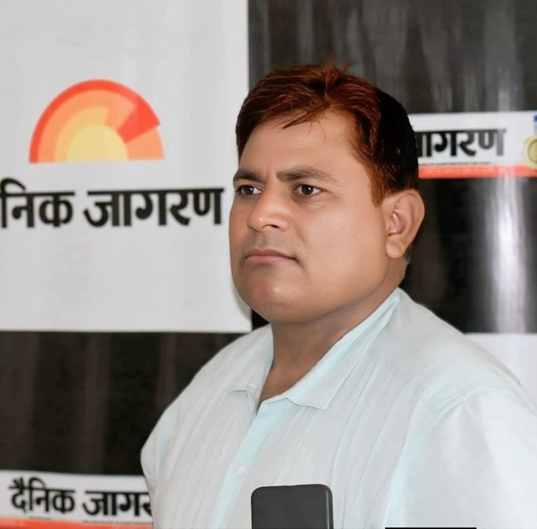 Portrait of a man in a light blue shirt standing in front of a Dainik Jagran banner with a sun logo in the background.
