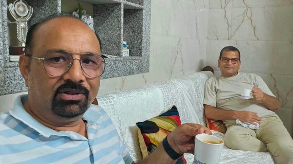 Two men sit on a white patterned couch indoors, each holding a mug of tea; foreground man wears a blue striped polo shirt and glasses, background man in beige shirt smiles nearby.