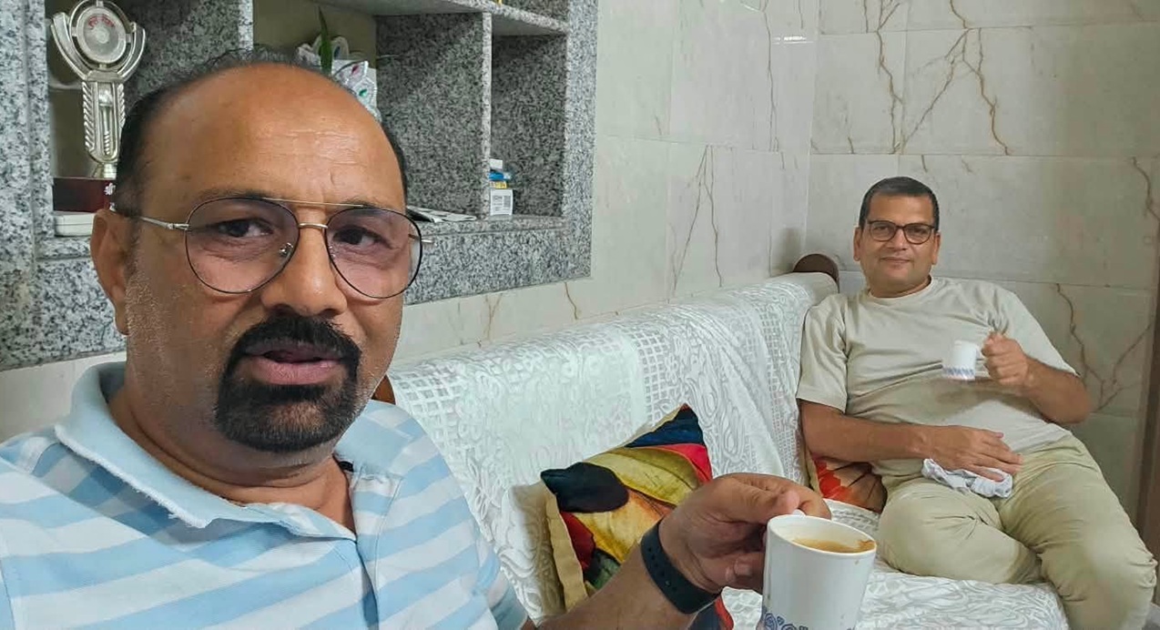 Two men sit on a white patterned couch indoors, each holding a mug of tea; foreground man wears a blue striped polo shirt and glasses, background man in beige shirt smiles nearby.