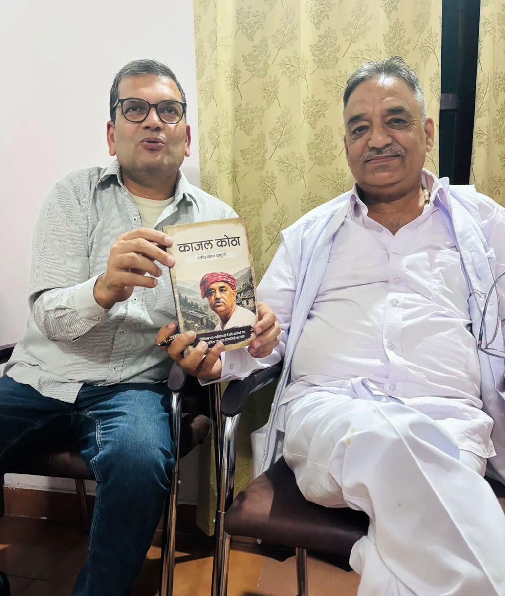 Two men sit side by side, one holding a book with a portrait on its cover visible to the camera.
