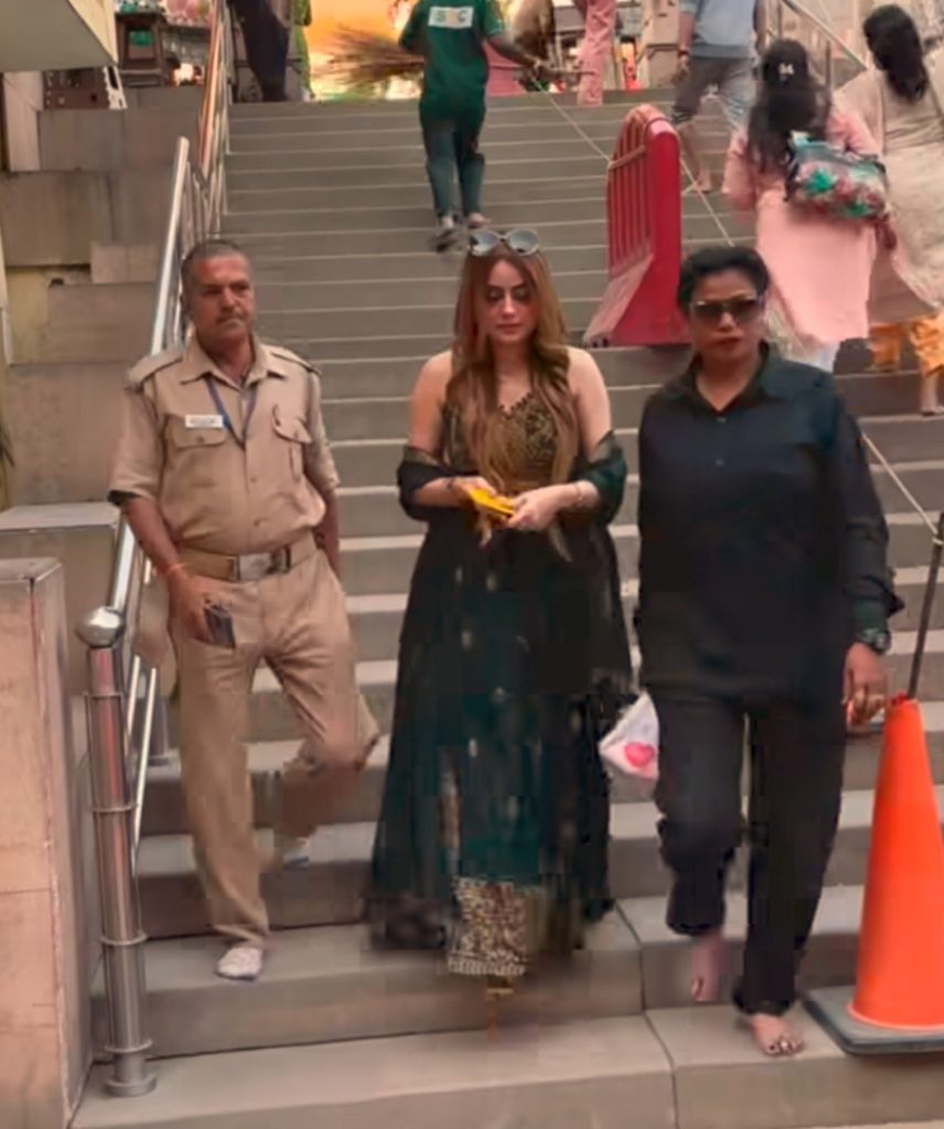 Three pedestrians descending outdoor stairs: a security guard in khaki uniform on the left, a woman in a leopard-print top with sunglasses on her head in the middle, and a woman in black with sunglasses on the right, with an orange traffic cone nearby.