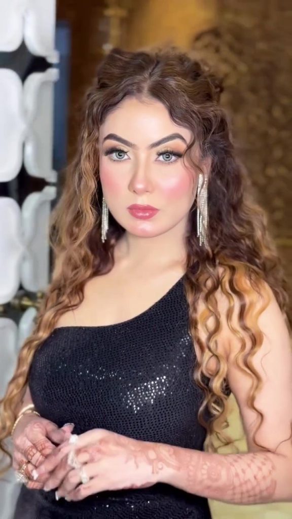 Portrait of a woman with long wavy hair, bold makeup, and diamond earrings wearing a black sequined dress, posed indoors.