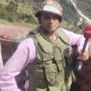 Male journalist wearing a PRESS helmet and tactical vest, holding a red NDTV microphone outdoors on a mountainous hillside.