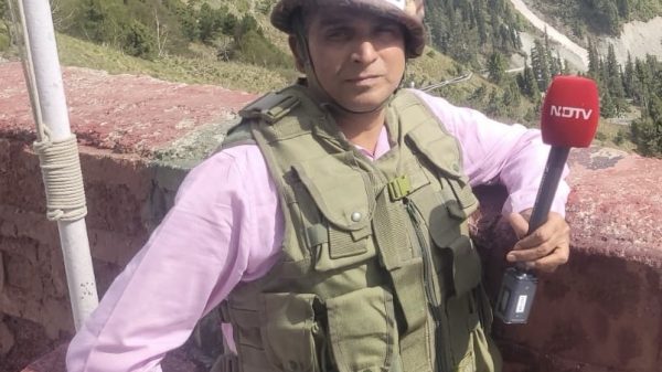 Male journalist wearing a PRESS helmet and tactical vest, holding a red NDTV microphone outdoors on a mountainous hillside.