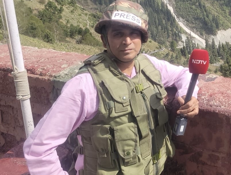 Male journalist wearing a PRESS helmet and tactical vest, holding a red NDTV microphone outdoors on a mountainous hillside.