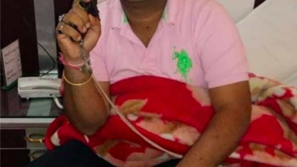 Man in a pink polo sits on a hospital bed, holding an IV line and smiling with a red blanket over his legs.