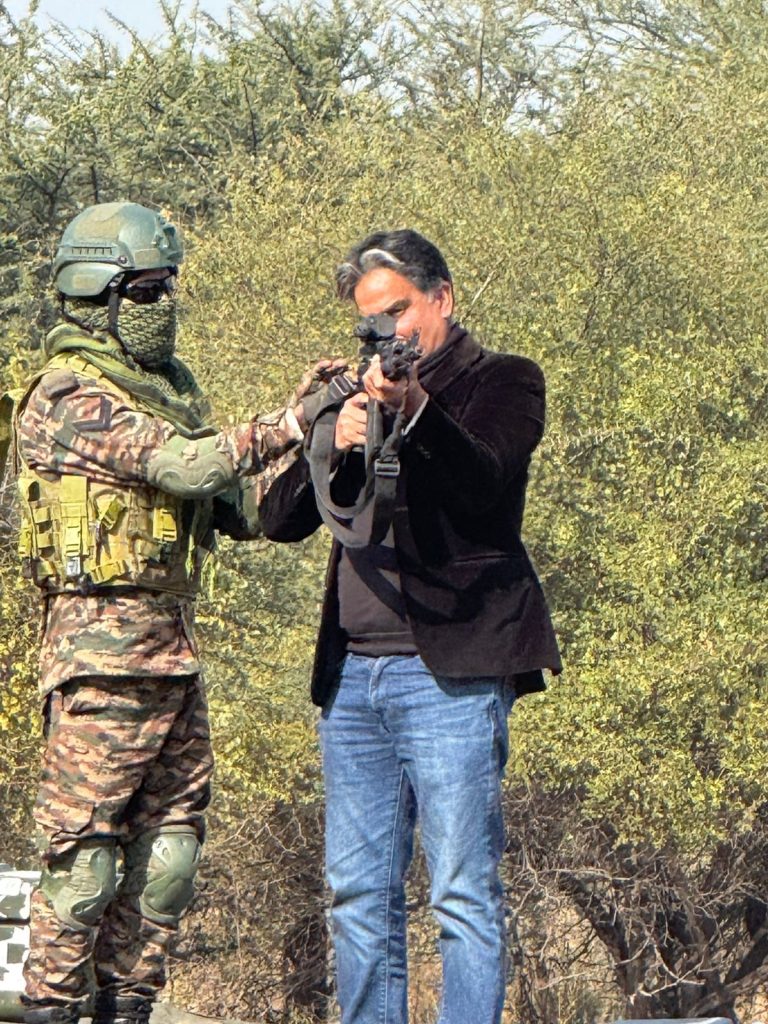 273d64d8-18a8-403b-9624-cdaabd13ab66 | No. 1 Indian Media News Portal Camouflage-clad soldier with helmet assisting a man in a dark jacket handling a rifle outdoors in a dry, bushy area.