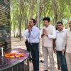 Group of adults standing in a tree-lined park, hands in prayer beside a black memorial plaque with gold inscriptions and a table covered in flower petals and a candle for a tribute.