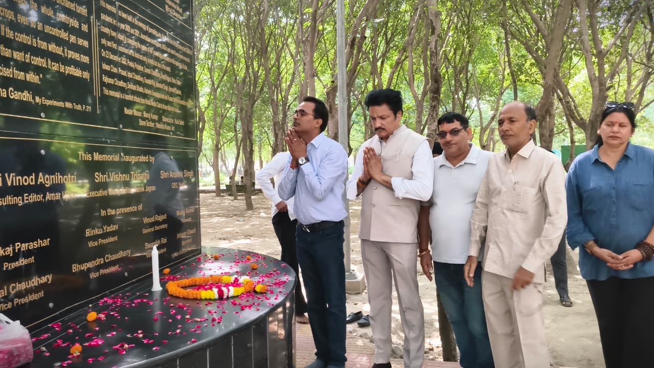 Group of adults standing in a tree-lined park, hands in prayer beside a black memorial plaque with gold inscriptions and a table covered in flower petals and a candle for a tribute.