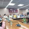 Group of men seated around a long conference table in a meeting room, microphones and water bottles in front of them, with a sign reading 'Indira Gandhi National Centre for the Arts' in the background.