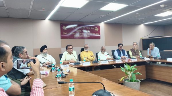 Group of men seated around a long conference table in a meeting room, microphones and water bottles in front of them, with a sign reading 'Indira Gandhi National Centre for the Arts' in the background.
