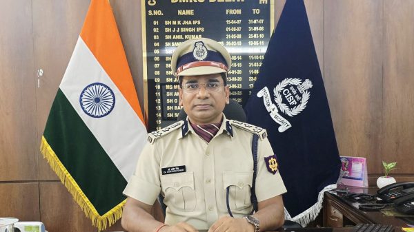 Indian security officer in uniform seated at a glass desk, with the Indian flag to his left and a CISF banner behind him, signing documents.