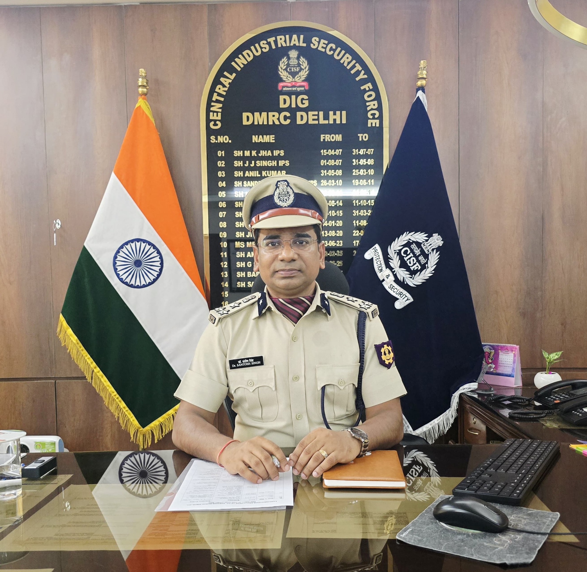 Indian security officer in uniform seated at a glass desk, with the Indian flag to his left and a CISF banner behind him, signing documents.