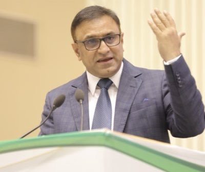 Man in a blue suit and glasses speaking at a podium with two microphones, gesturing with his right hand.