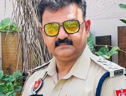 Male police officer in khaki uniform with medals and yellow-tinted sunglasses, standing outdoors.